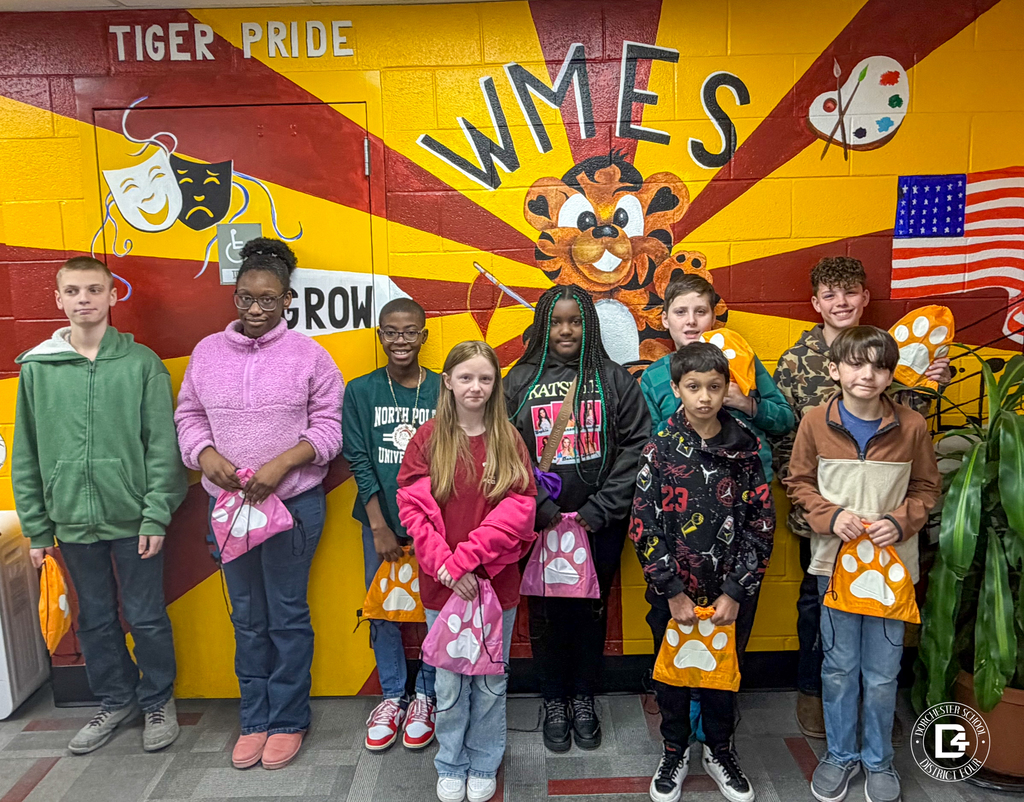 A group of upper elementary and middle school students pose in front of a “WMES” tiger mural, holding colorful paw-print drawstring bags and smiling at the camera.