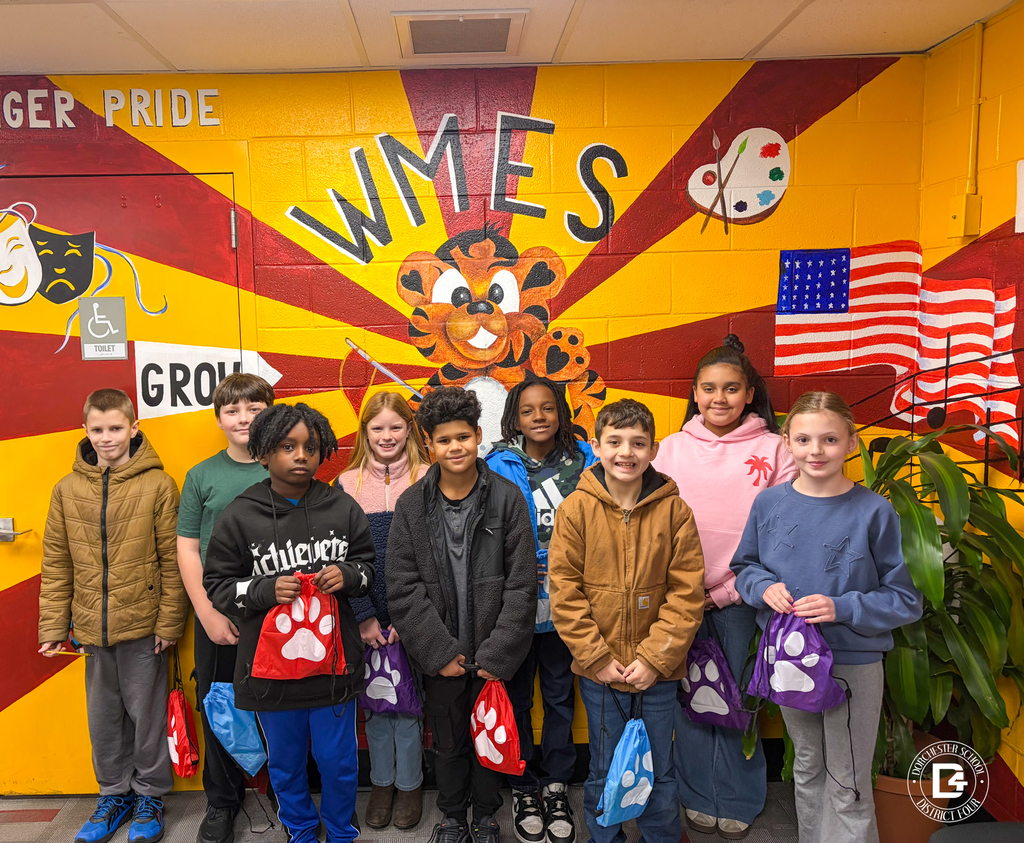 A class of elementary students stands shoulder to shoulder in front of the WMES tiger mural, each holding a paw-print drawstring bag, with bright school colors filling the background.