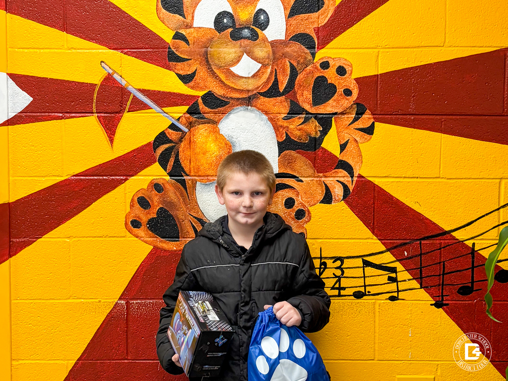 A single elementary-aged student stands in front of the WMES tiger mural holding a boxed prize and a blue paw-print drawstring bag.