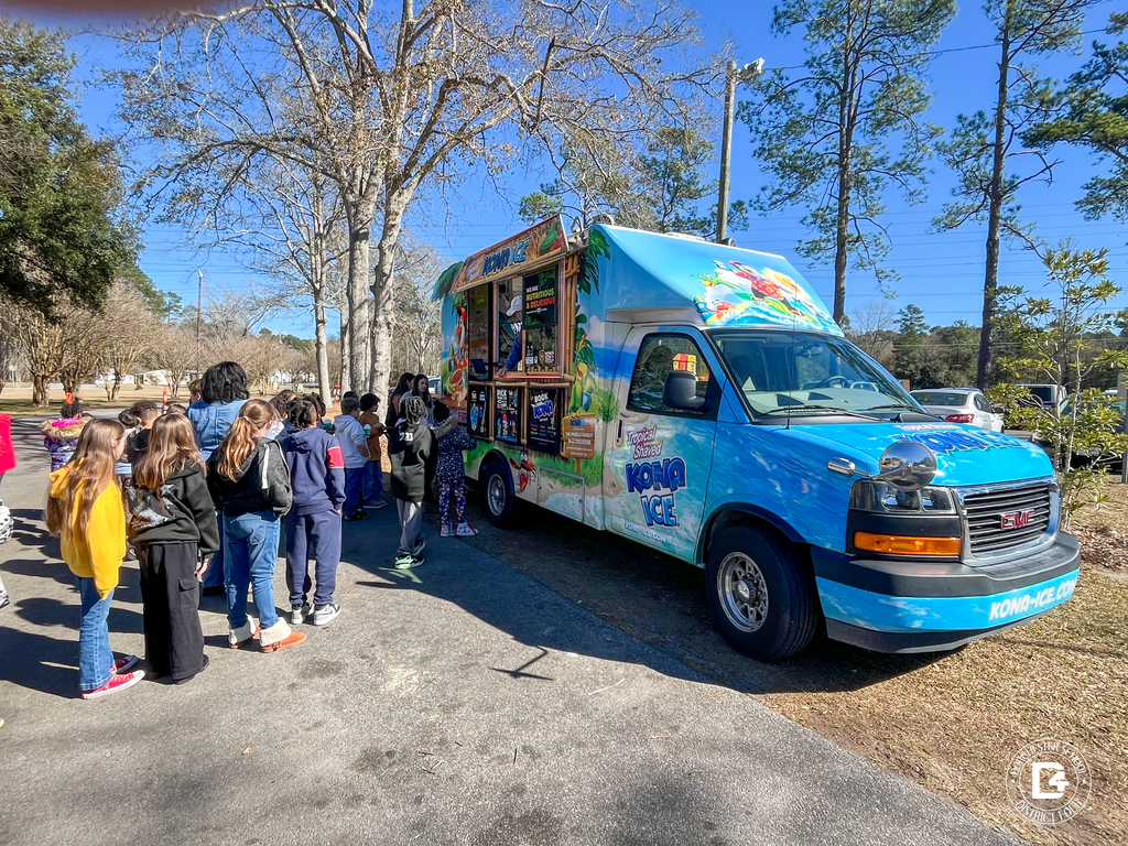 A Kona Ice truck is parked outside on a sunny day as a long line of elementary students waits to order shaved ice.