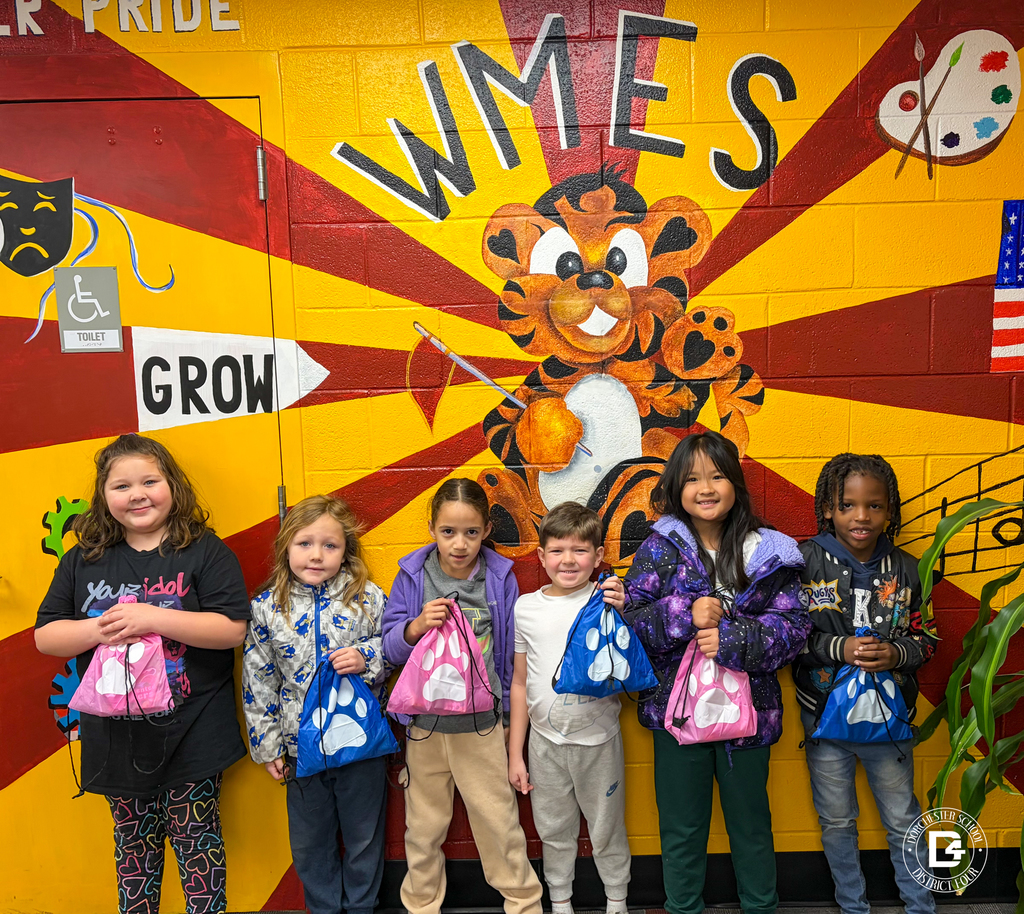A group of younger elementary students stand in front of the WMES tiger mural, holding blue and pink paw-print drawstring bags.