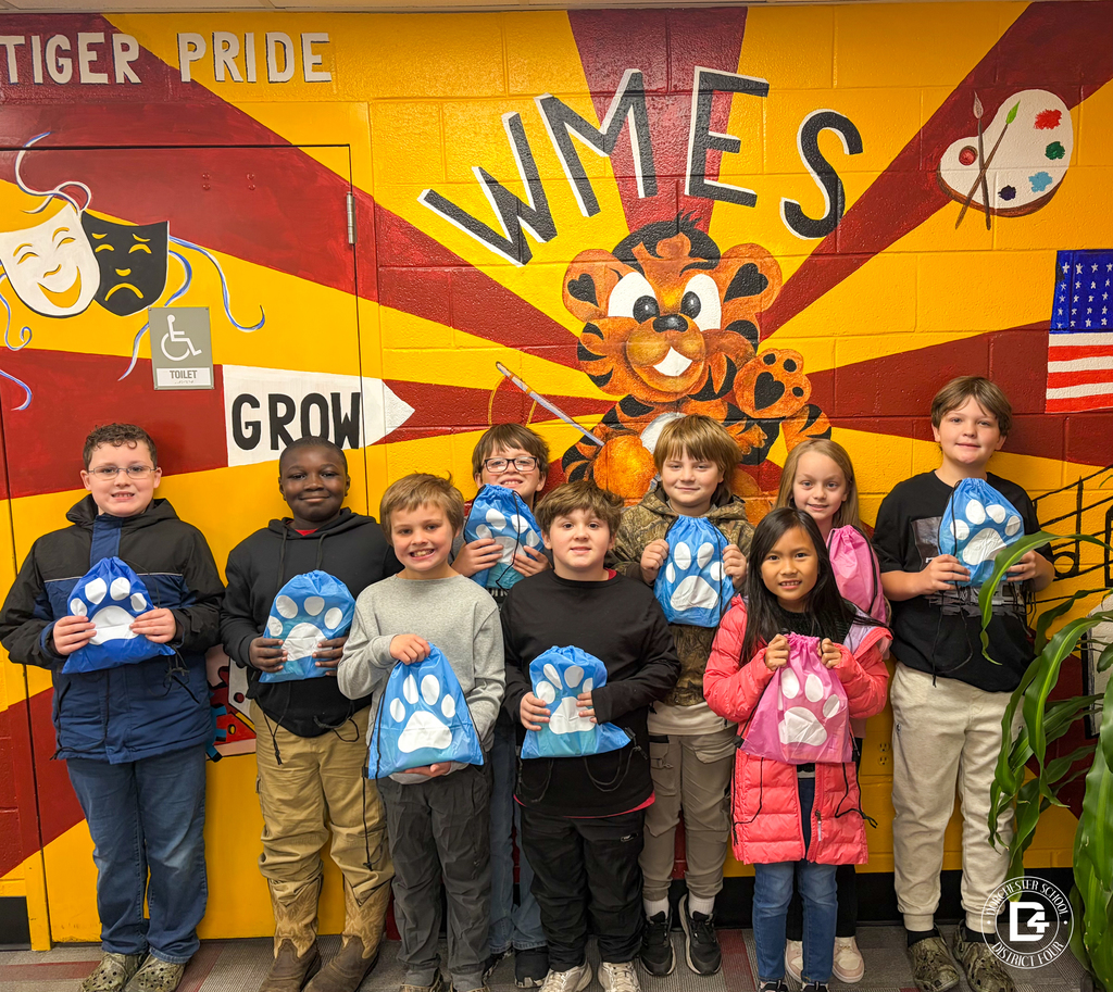 A group of elementary students pose in front of the WMES tiger mural, holding blue and pink paw-print drawstring bags, with the Dorchester School District Four logo visible in the corner.