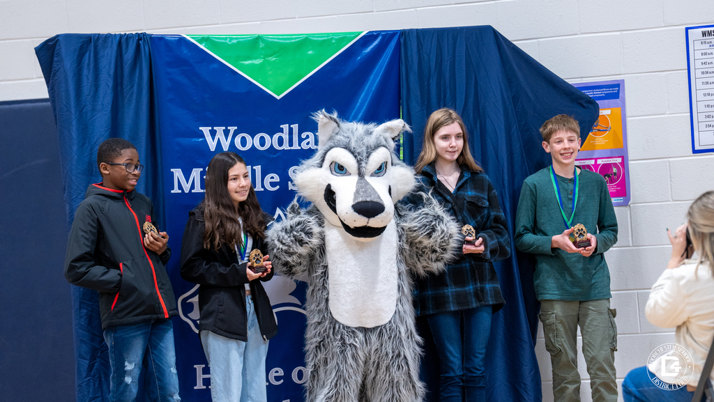 A group of Woodland Middle School students stand with the school mascot holding academic awards in front of a Woodland Middle School Wolves backdrop.