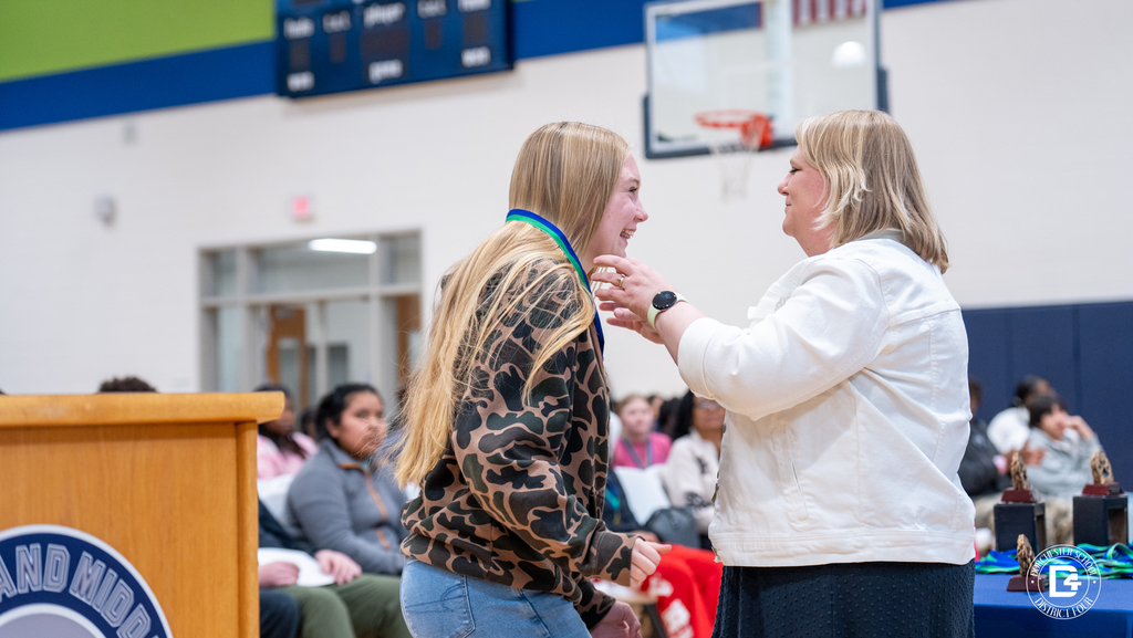 A Woodland Middle School staff member places a medal around a smiling student’s neck during the Quarter 2 awards ceremony with students seated in the background.