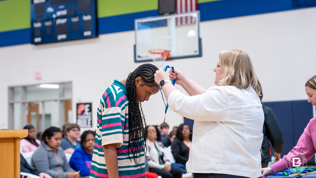A Woodland Middle School staff member places a medal over a student’s head during the Quarter 2 awards ceremony while other students watch.