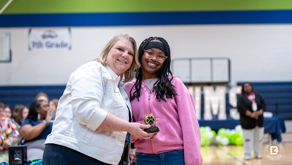 A Woodland Middle School staff member and student pose together holding an academic award as the audience applauds during the Quarter 2 ceremony.