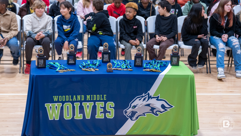 A table displaying trophies and medals for the Woodland Middle School Quarter 2 Awards is set up in the gym with students seated behind it.
