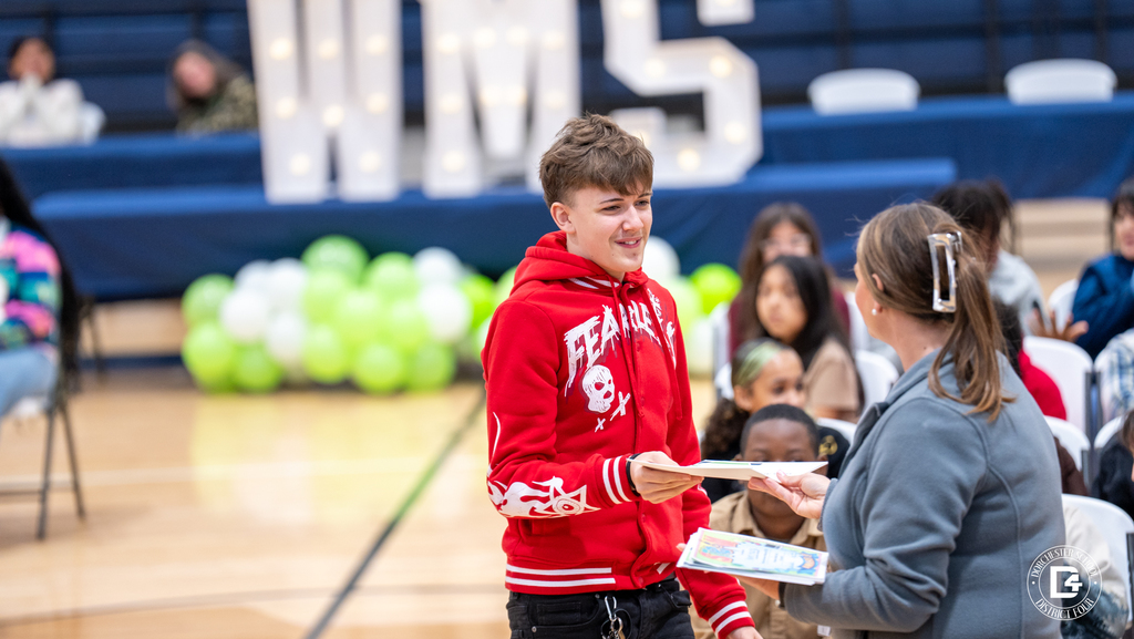 A Woodland Middle School student in a red hoodie smiles while receiving a certificate from a staff member as other students look on during the Quarter 2 awards ceremony.