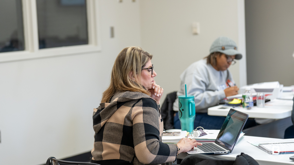 A teacher works on a laptop during DD4 Teacher Mentor Training while another participant takes notes nearby.