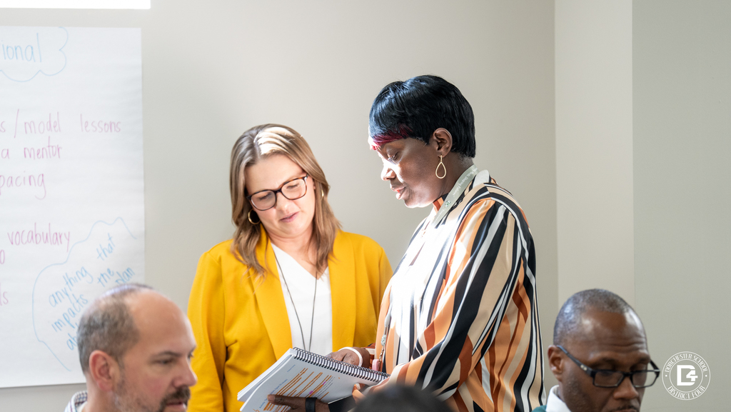 Two educators stand together reviewing mentor training materials while other participants work at tables nearby.