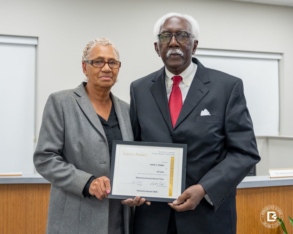 Dr. James Hodges stands with his wife while holding a framed Service Award recognizing 35 years of service during a DD4 board meeting.