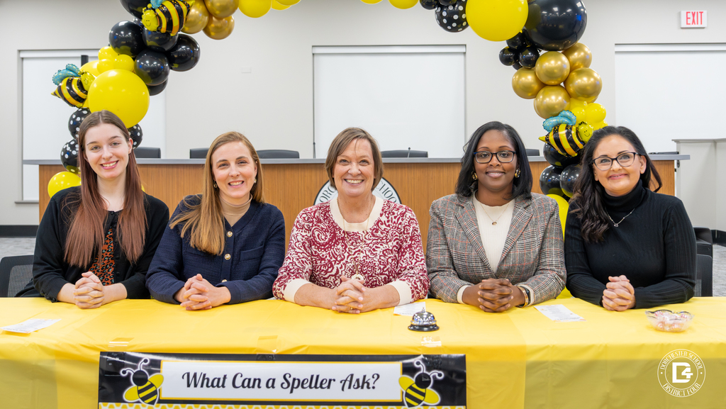 Five adult spelling bee officials sit side by side behind a yellow table labeled “What Can a Speller Ask?” inside a meeting room, framed by black, gold, and yellow balloon arches with bee decorations, serving as judges, recorder, and pronouncer for the Dorchester School District Four spelling bee.