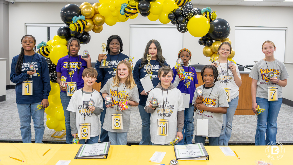 Group photo of elementary and middle school students standing in two rows indoors beneath a black, gold, and yellow balloon arch with bee decorations, each holding spelling bee trophies or awards, with a table in front displaying certificates and programs during the Dorchester School District Four spelling bee event.