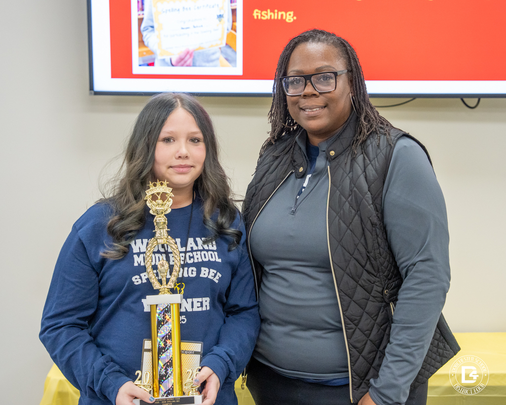 The spelling bee champion, a middle school student from Woodland Middle School, stands indoors holding a large trophy beside an adult staff member, following the Dorchester School District Four spelling bee.