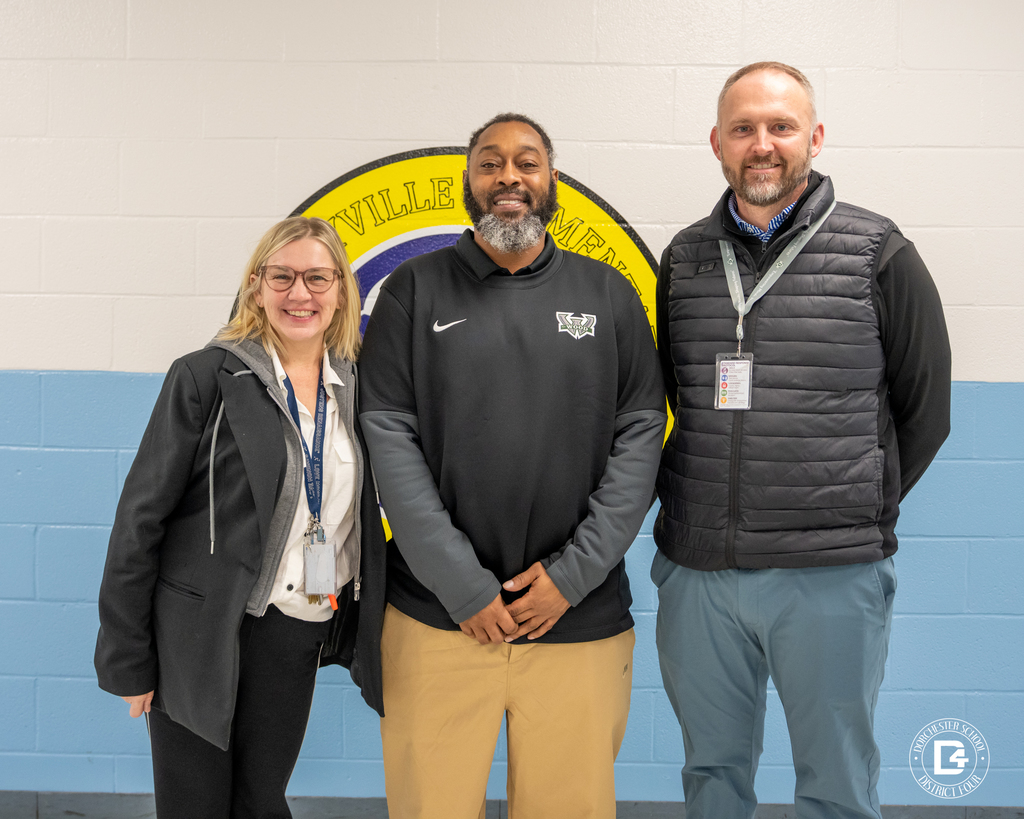 Three adults pose in front of a school wall emblem during a Dorchester School District Four Aspiring Administrator Program visit.