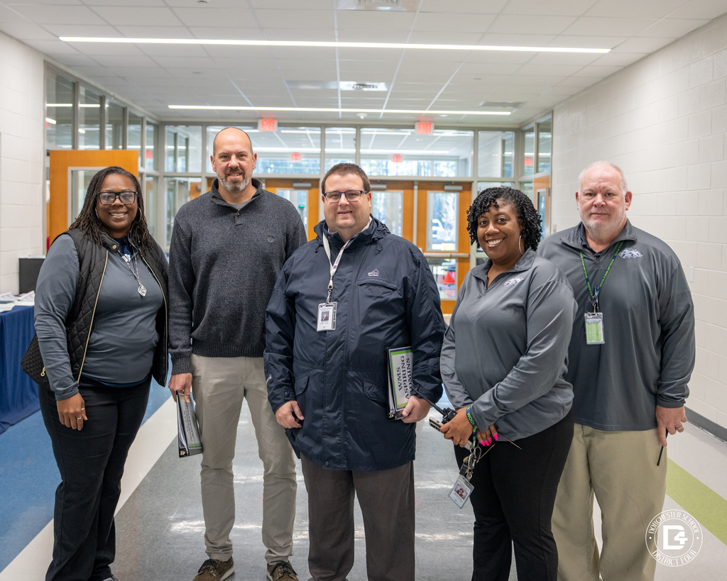 Five adults stand together in a bright school hallway with glass doors behind them, posing during an Aspiring Administrator shadowing visit.