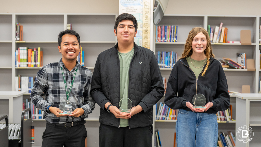 two Woodland High School students and a teacher pose in the media center holding Wolverine of the Month awards, with bookshelves behind them.
