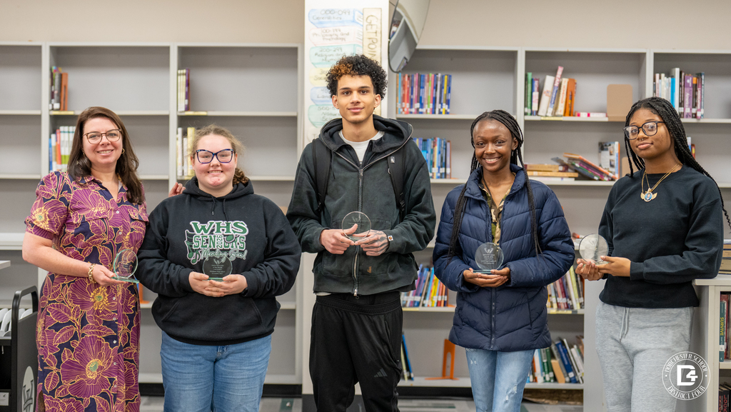Six Woodland High School Wolverines of the Month, including students and a staff member, stand together holding awards in the media center.