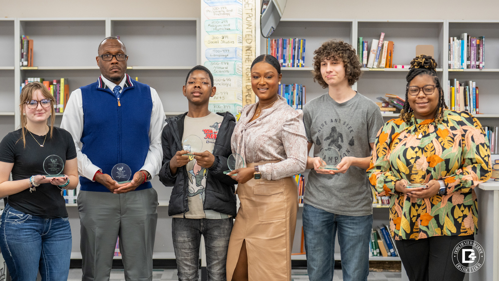 Seven Woodland High School Wolverines of the Month, including students and staff, stand in the media center holding glass recognition awards.
