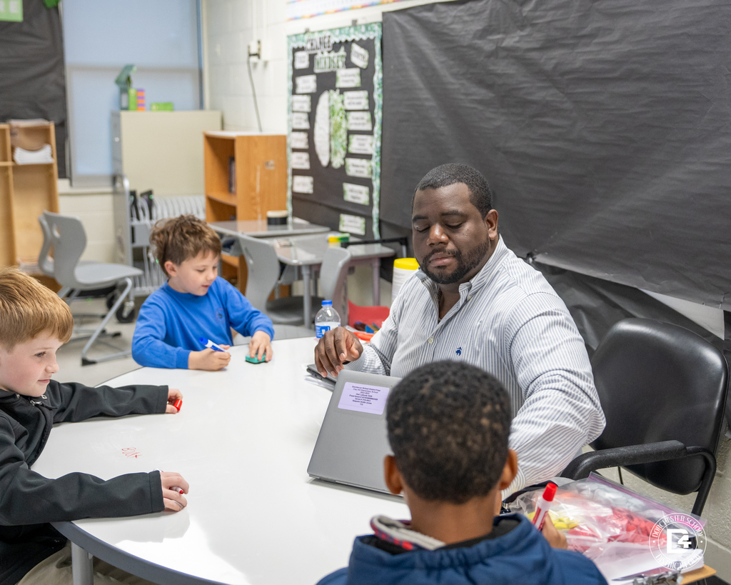 A teacher works with several elementary students at a round table, handing out materials as students write with dry-erase markers during small group instruction.