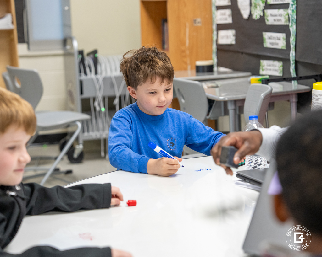 An elementary student sits at a desk working independently on a Chromebook, focused on the screen with classroom materials and posters visible in the background.