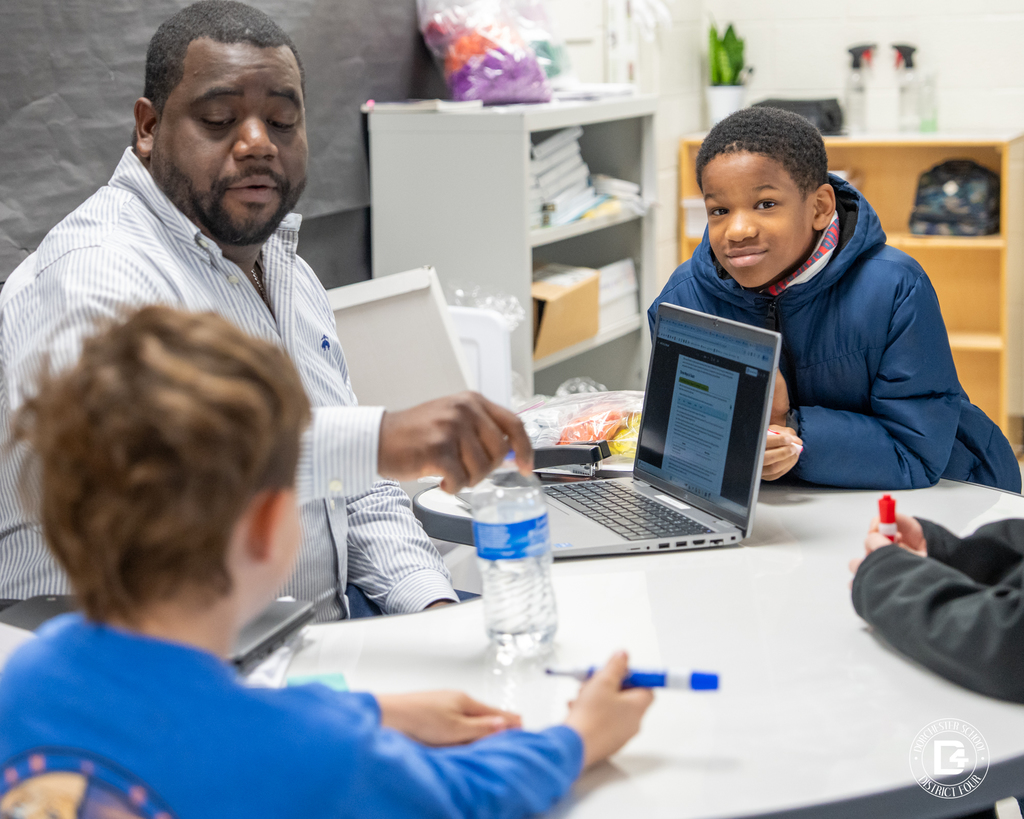 A teacher points toward materials on a table as students sit nearby, one student looking toward the camera while others follow along during a small group learning activity.