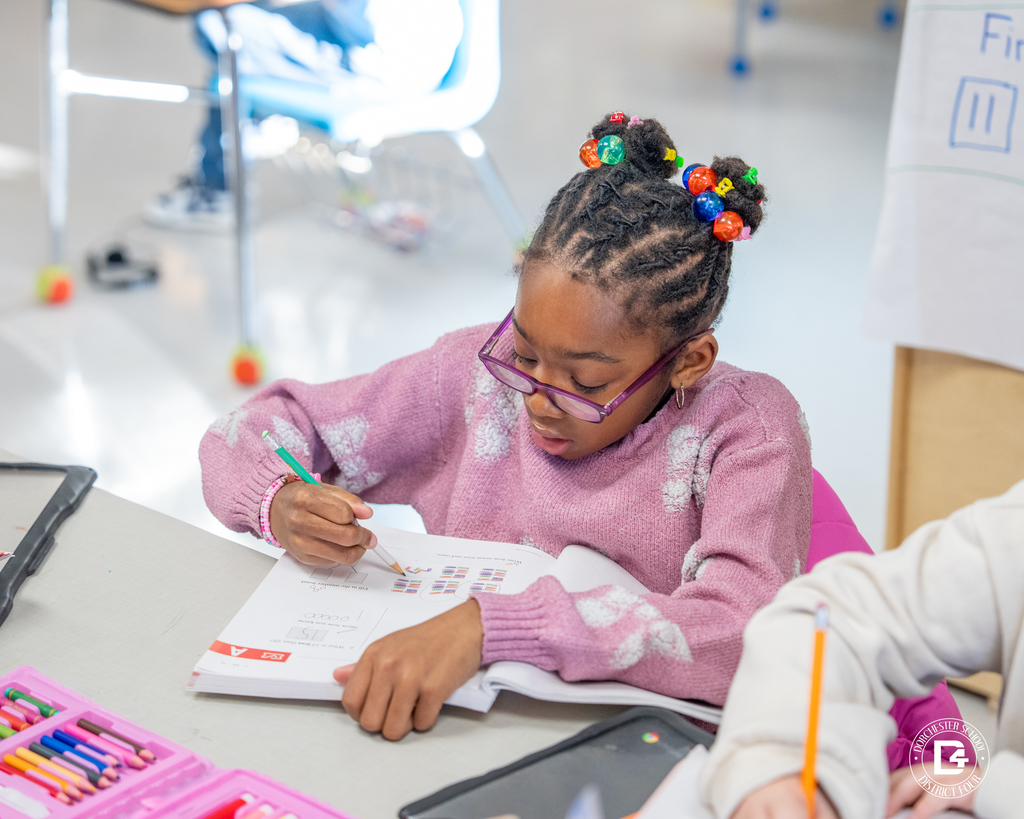 A student with glasses works carefully in a math workbook at a classroom table, coloring and writing answers.