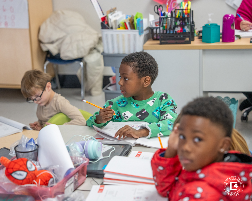 A first-grade student looks up from a workbook holding a pencil, with classmates working nearby.