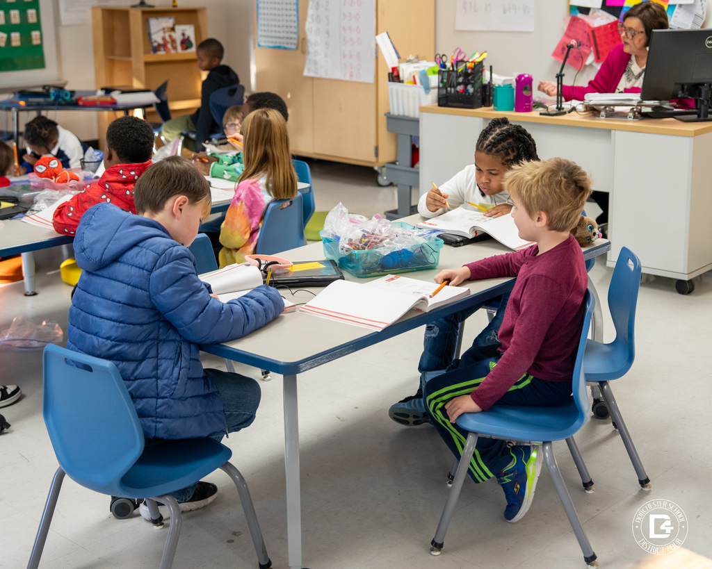 Several first-grade students sit at grouped tables, each working independently in their math books during class.