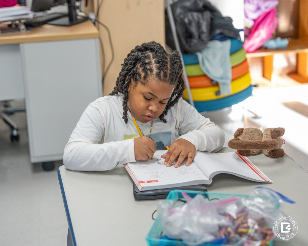 A first-grade student sits at a table, focused on completing a math worksheet in a workbook using a pencil.