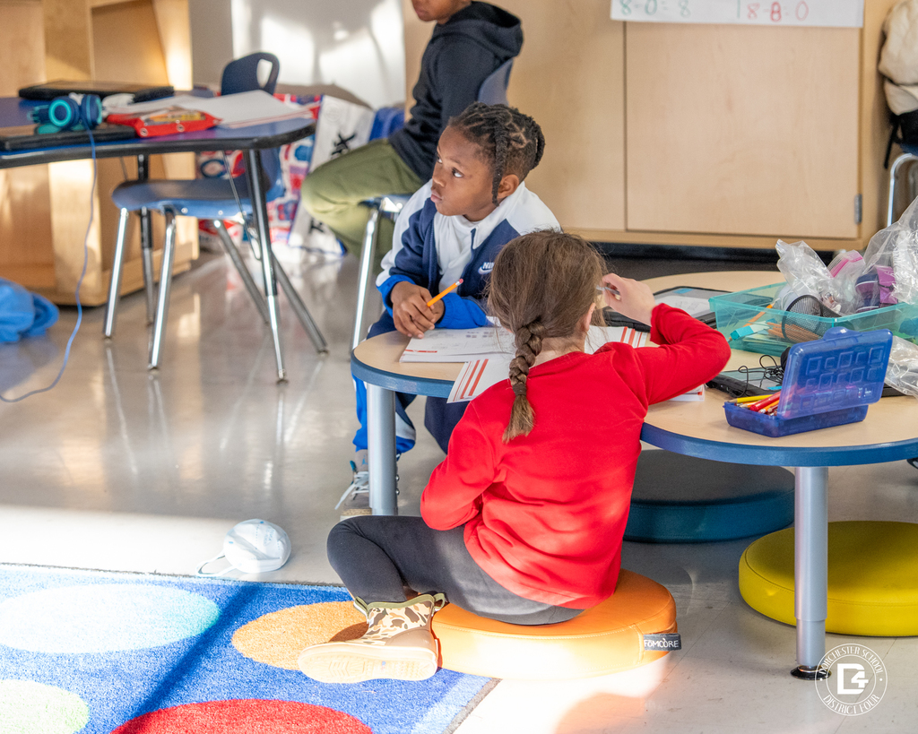 A student sits on a round classroom seat at a low table, writing in a workbook while sunlight fills the room.