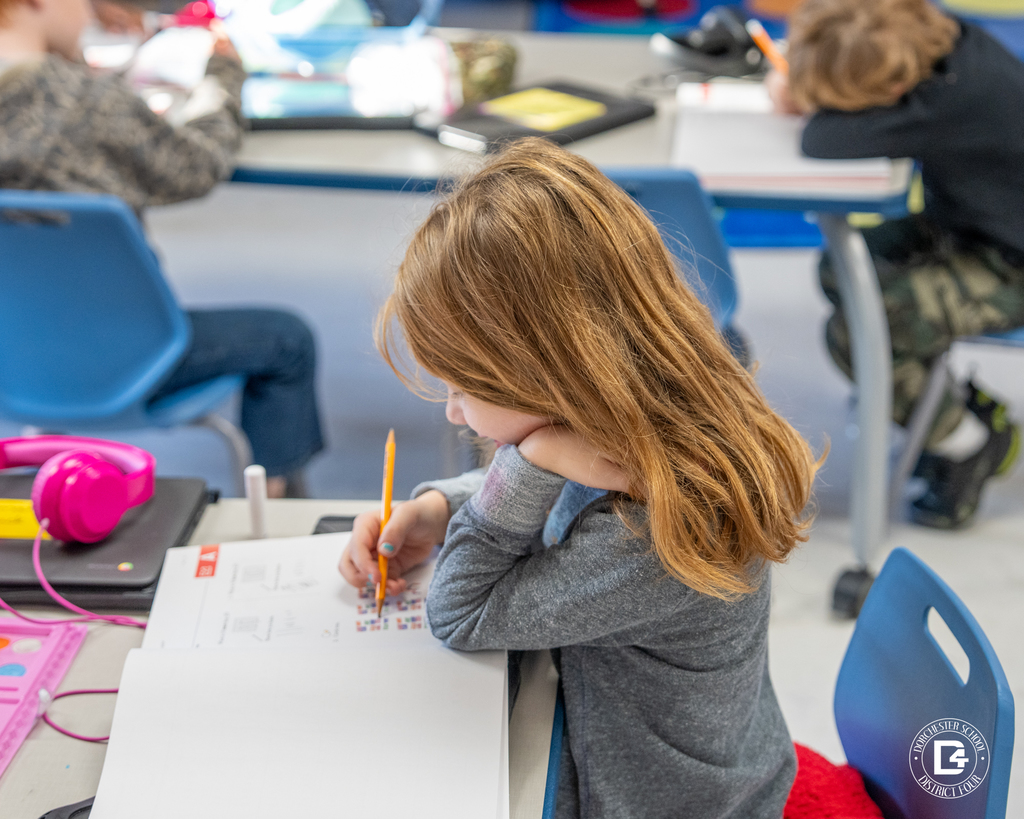 A first-grade student sits at a table, focused on completing a math worksheet in a workbook using a pencil.
