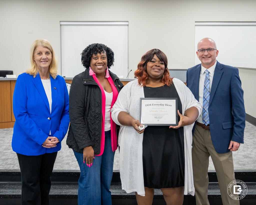 Four adults stand indoors at a school board meeting. Mrs. Latisha Brown holds a DD4 Everyday Hero certificate and coin while standing with district leaders in front of a meeting room backdrop.