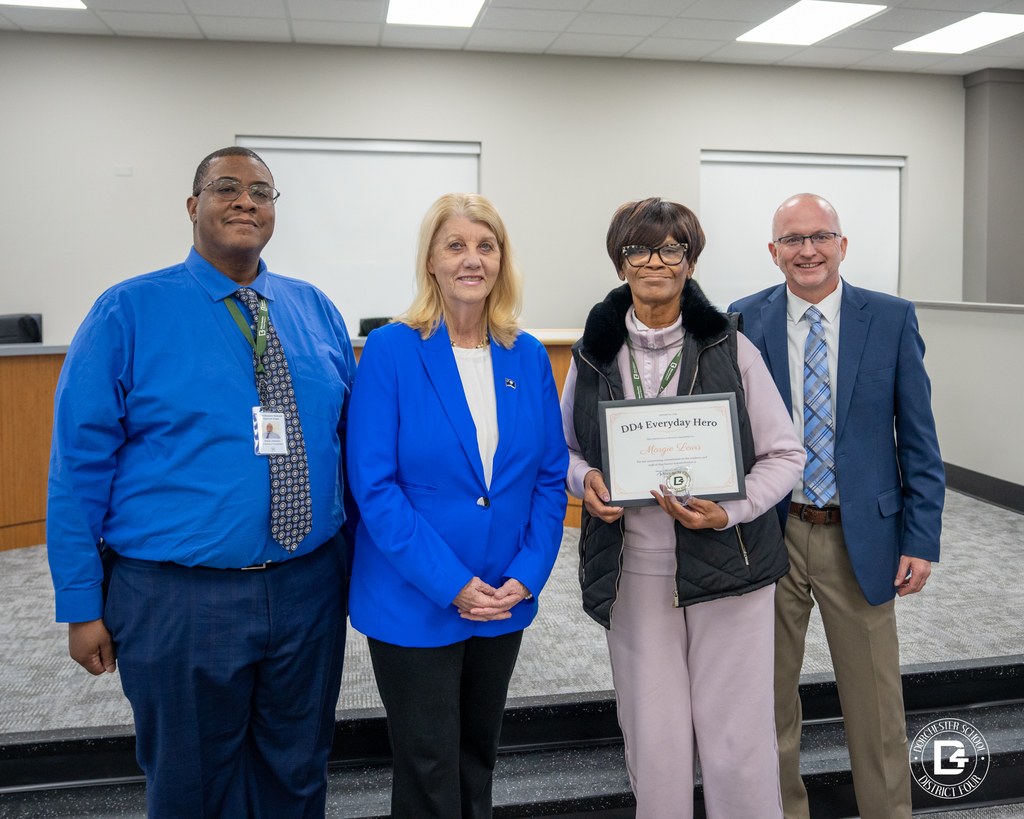 Four adults pose at a school board meeting. Mrs. Margie Lewis holds a DD4 Everyday Hero certificate and coin, standing beside district leaders in a meeting room.