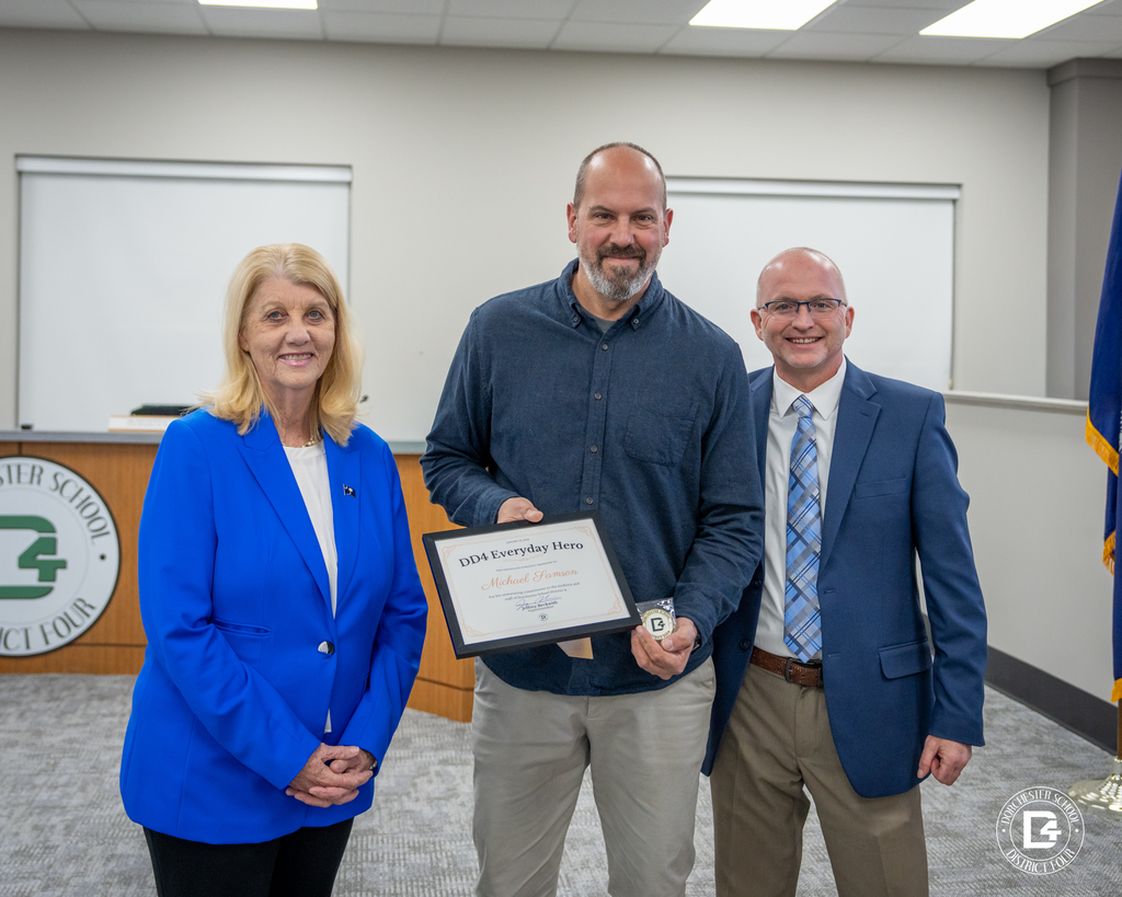 hree adults stand at a school board meeting. Mr. Michael Samson holds a DD4 Everyday Hero certificate and coin while standing with district leaders near the board dais.