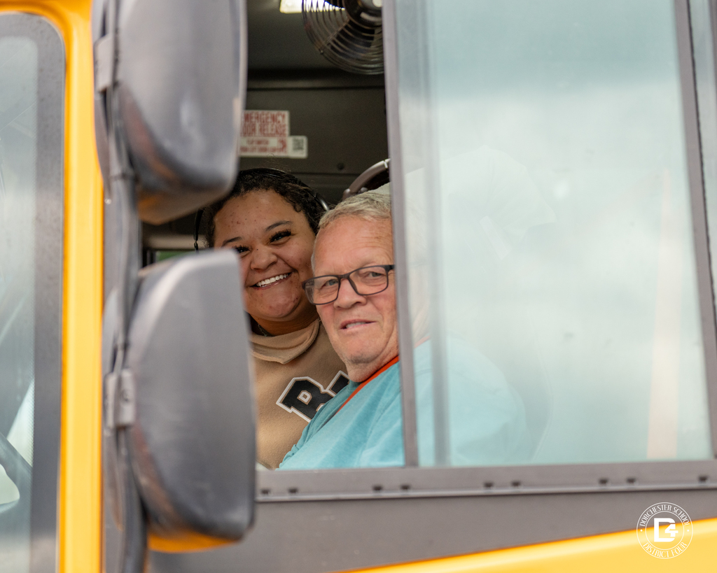 Two school transportation staff members smile from inside a yellow school bus, visible through the open window.