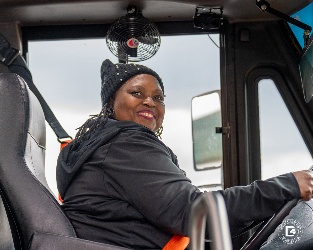 School bus driver wearing a black jacket and knit hat smiles while seated behind the wheel, interior fan visible above.