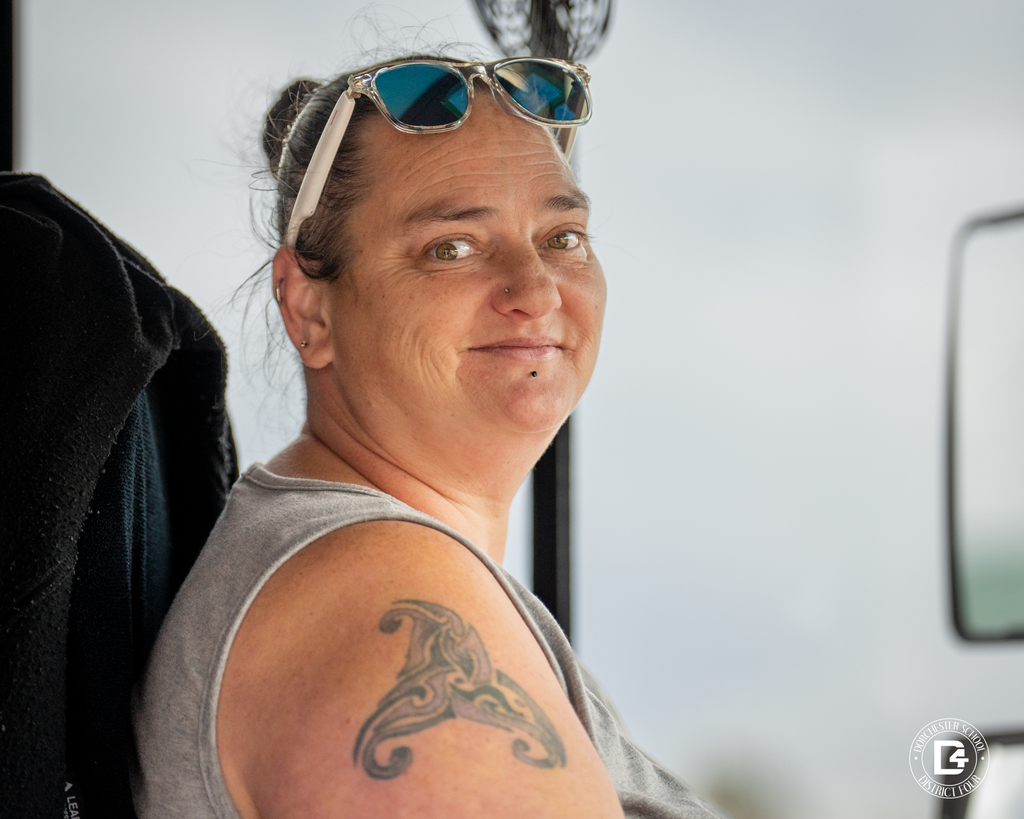 School bus driver with sunglasses on her head and visible arm tattoo looks toward the camera from the driver’s seat.