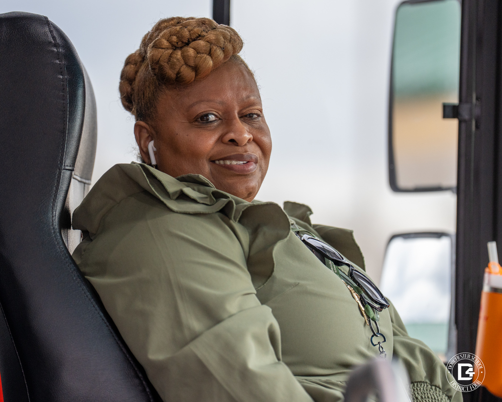 School bus driver wearing a green blouse sits in the driver’s seat, looking toward the camera.