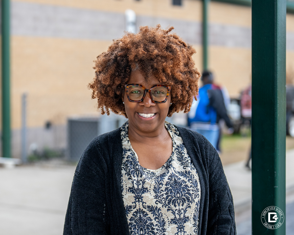 Smiling woman with curly hair and glasses stands outside on a school campus near green poles, wearing a patterned blouse and black cardigan.