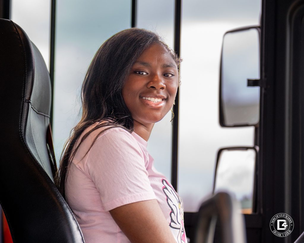 Young woman seated in the driver’s seat of a school bus, smiling toward the camera with hands near the steering wheel.