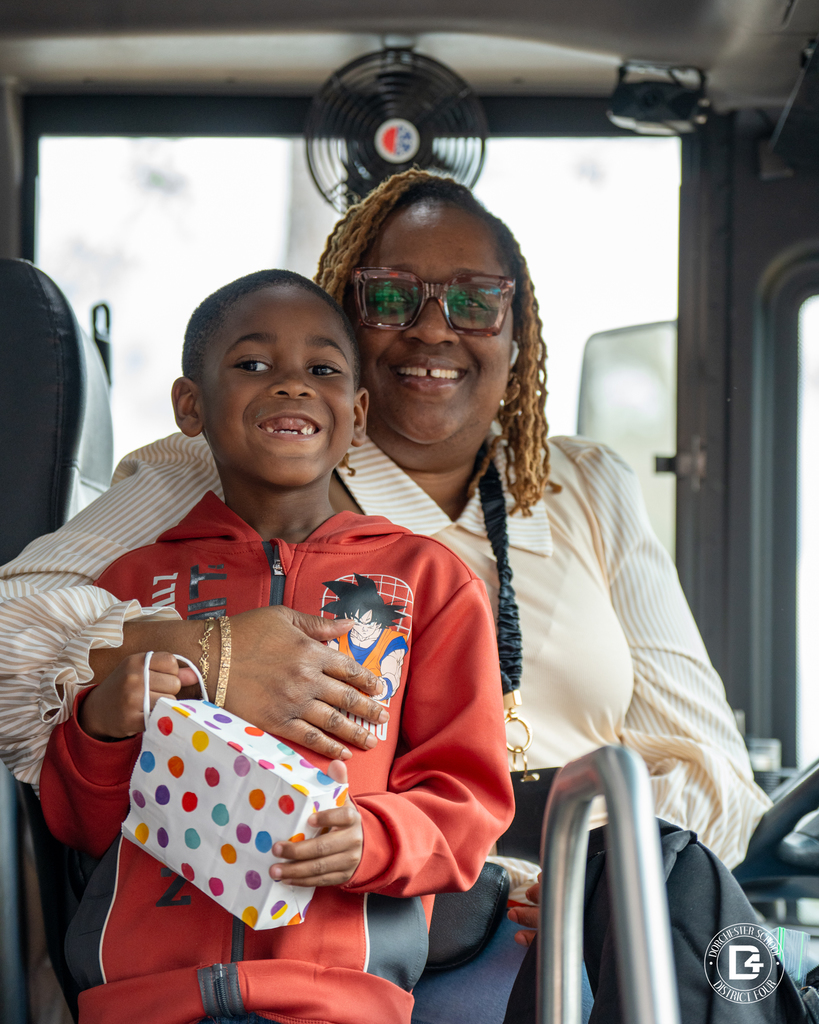 School bus driver smiles while holding a young student seated on her lap inside the bus, the child holding a small gift bag.
