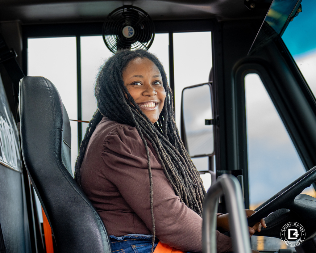 School bus driver with long locs smiles while seated behind the wheel inside the bus.