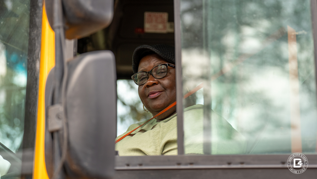 A smiling school bus driver wearing glasses and a dark cap looks out from the driver’s seat through the open bus window, seatbelt fastened, with the yellow exterior of the bus visible along the frame.