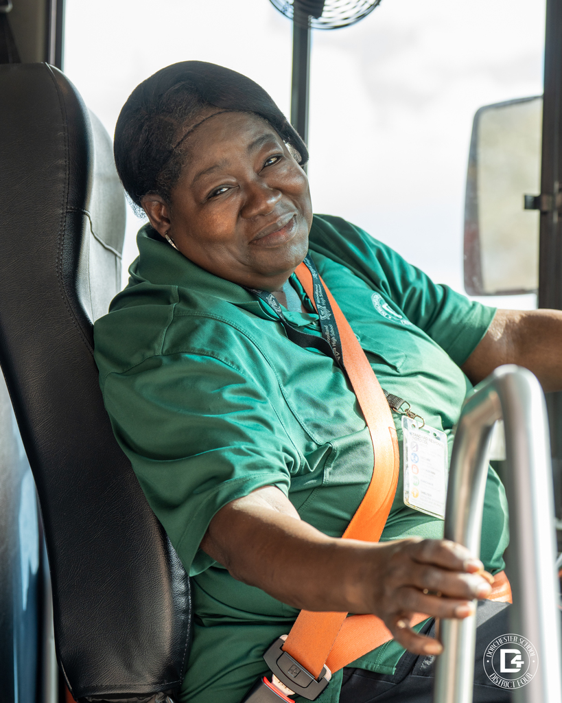 A school bus driver wearing a green transportation polo and orange safety belt sits in the driver’s seat, smiling warmly while holding the steering wheel inside the bus.