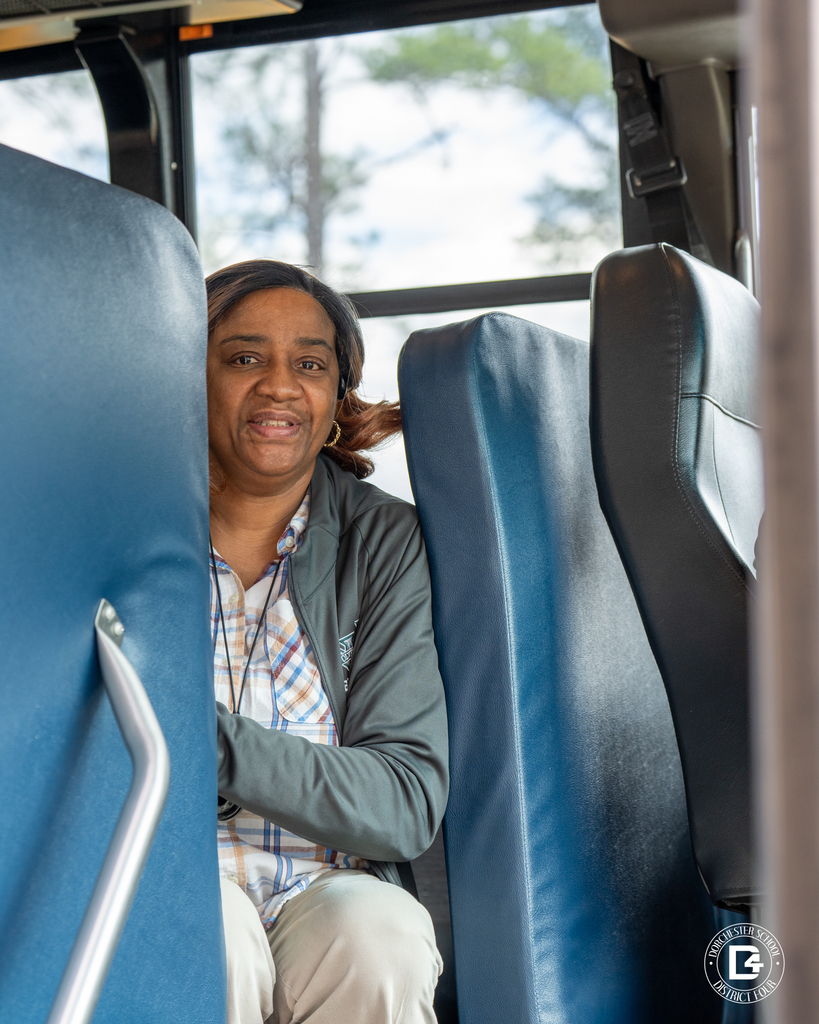 A bus driver sits between blue bus seats inside the vehicle, wearing a plaid shirt and jacket, looking toward the camera with a calm expression.