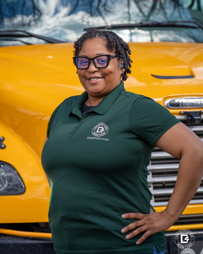 A transportation staff member wearing a green Dorchester School District Four polo stands with one hand on her hip in front of a yellow school bus grille, smiling at the camera.