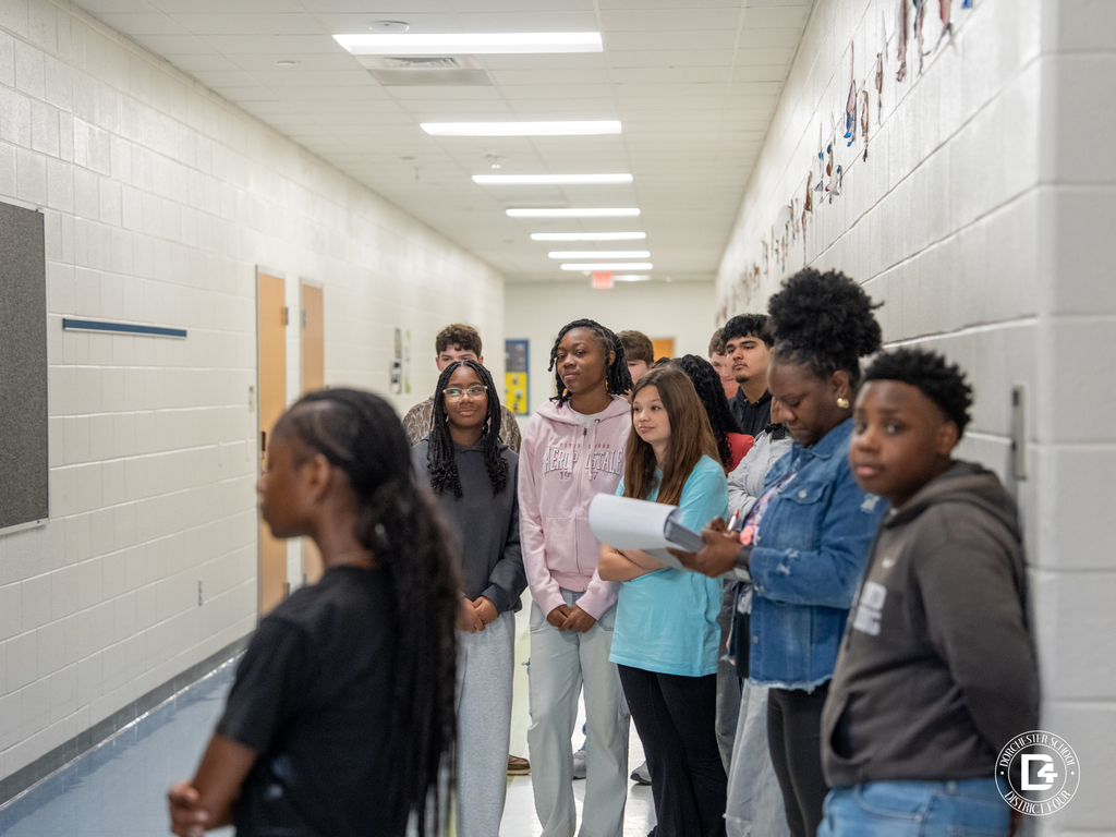 A group of students stands together in the hallway, listening and observing as a classmate presents information from a Black History Month project.