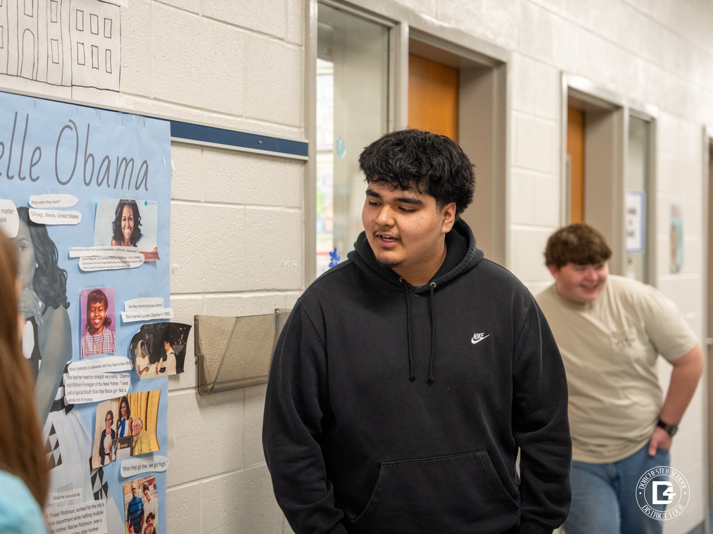 A student stands in the hallway presenting a door display about Michelle Obama, pointing toward photos and printed research while another student listens nearby.