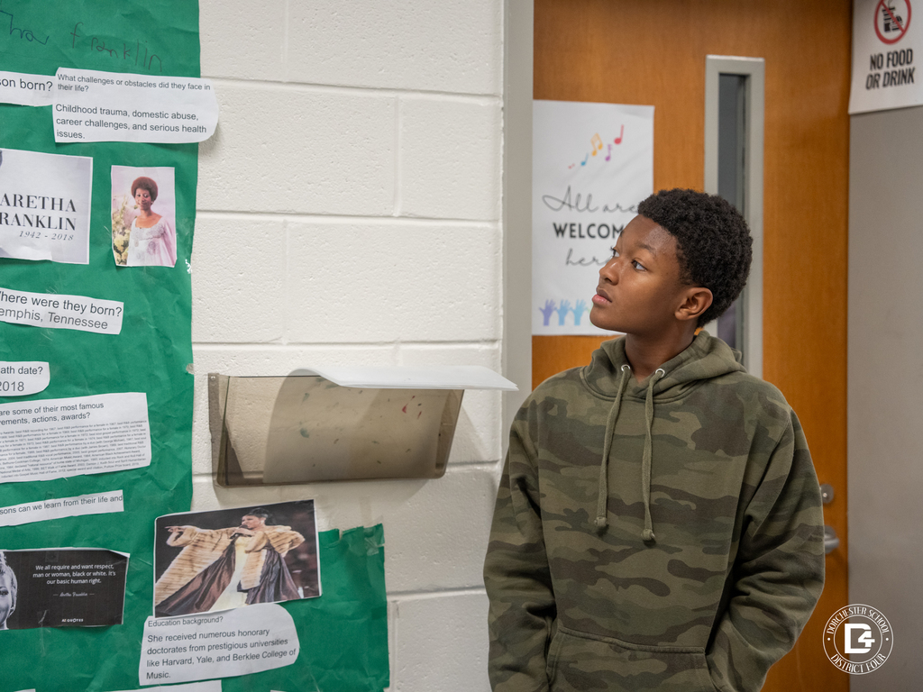 A student looks up at a green poster about Aretha Franklin, reading printed facts about her life, accomplishments, and legacy.